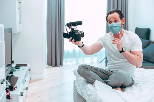 의료용 마스크를 침대에 카메라에 자신을 기록하는 손가락을 가리키는 젊은이 — 스톡 사진