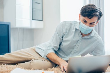 Sağlık maskeli genç beyaz adam pantolonlu, gömlekli ve dizüstü bilgisayarının klavyesinde daktilo kullanıyor.