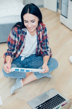 Gömlekli ve kotlu neşeli bayan evde yerde oturuyor ve elinde grafik çizelgesine bakarken gülümsüyor.