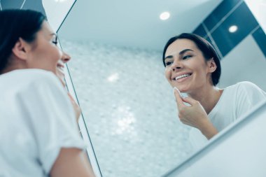 Memnun genç bayan gülümsüyor ve aynaya bakıyor ve yüzünü pamukla temizliyor.