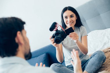 Neşeli siyah saçlı kadın, gülümseyen ve önünde duran adama mikrofonlu kamerayla bakıyor.