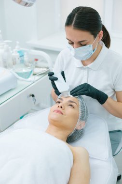 Konsantre bir kozmetik uzmanı alnında ultrasonik temizleme işlemi yaparken neşeli bir kadın gülümsüyor.