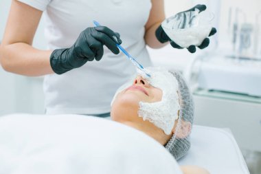 Güzellik kliniğinde yatarken çekilmiş bir fotoğraf ve plastik eldivenli kozmetik uzmanının elleri yüzüne aljinat maske seriyor.