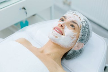 Banyo şapkalı beyaz bir kadının portresi tıbbi bir kanepede uzanmış ve yüzüne oksijen maskesi takmış gülümsüyor.