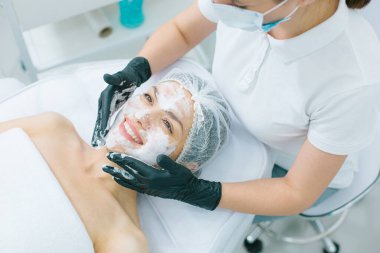 Güzellik salonunda yatan mutlu bir kadın ve plastik eldivenli profesyonel bir kozmetikçi yüzüne oksijen maskesi sürüyor.