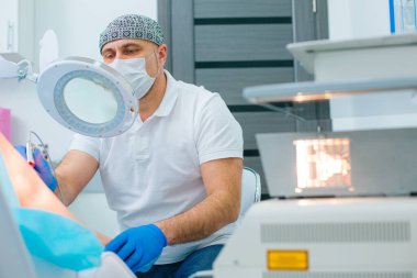 Tıbbi maskeli ciddi bir dermatolog modern klinikte lazer örümcek damarlarını temizliyor.