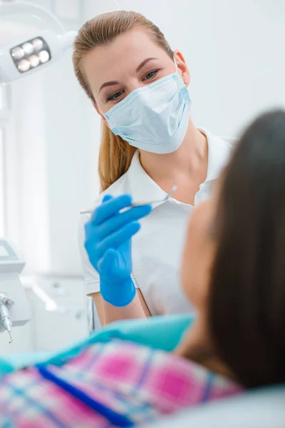 Female doctor wearing medical mask holding a mouth mirror while standing in front of her patient