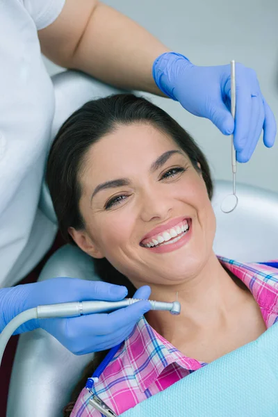 stock image Dentist in blue rubber gloves holding a high speed handpiece and a mouth mirror near the face of a positive smiling woman