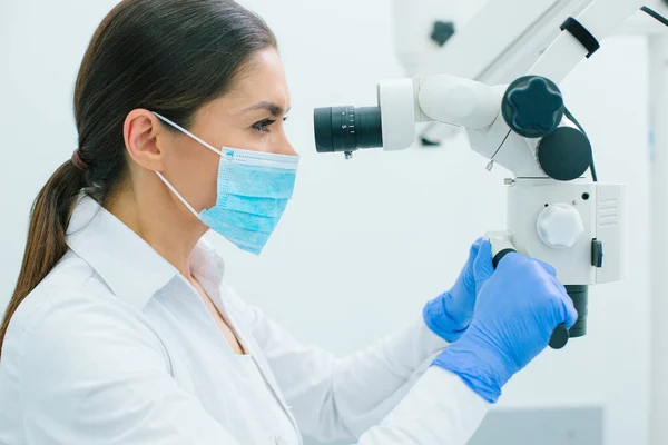 Female medical specialist in rubber gloves and mask putting hands on the handles of a microscope while working