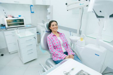 Mirthful Caucasian woman in casual clothes sitting in a dental chair of a modern office and smiling clipart