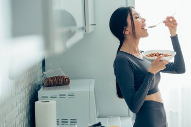 Güzel genç bayan mutfakta elinde bir kase mısır gevreğiyle duruyor ve ağzına bir kaşık sokuyor.
