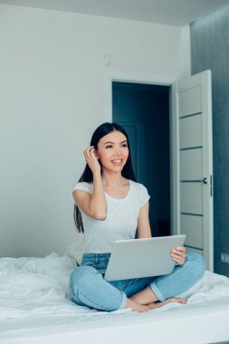 Güzel beyaz kadın saçlarına dokunuyor ve dizüstü bilgisayarla yatakta otururken gözlerini kaçırıyor.