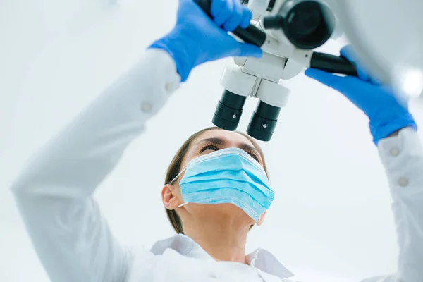 Concentrated dentist in medical mask and rubber gloves looking through the lenses of a surgical microscope and touching the handles