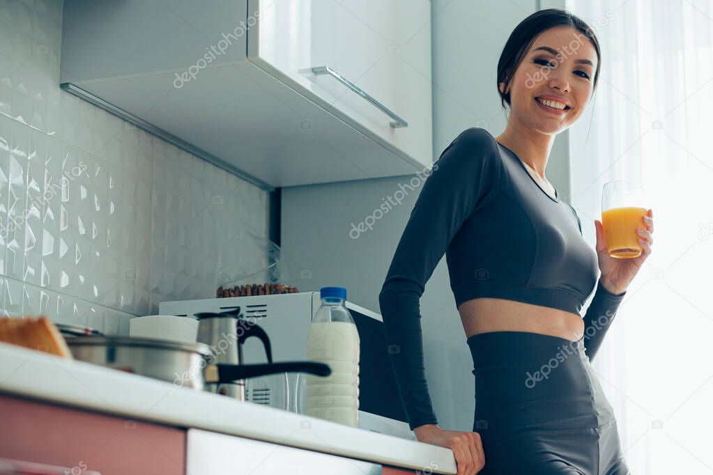 Confident pretty lady in sports clothes standing with a glass of orange juice in the kitchen. Website banner