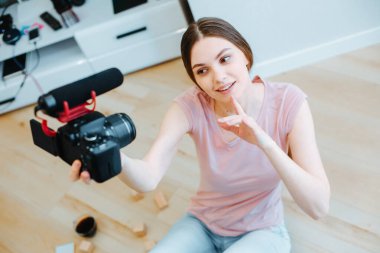 Kafkasyalı bir kadın, gündelik giysiler içinde, elinde kamera ve mikrofonla evde oturuyor.