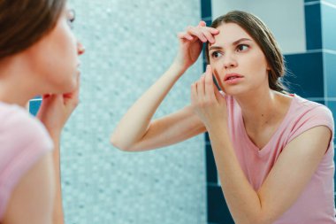 Ciddi genç bayan aynaya bakarken yüzüne ve gözünün etrafındaki deriye dikkatlice dokunuyor.