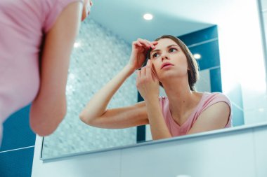 Ciddi bir genç kadın banyo aynasındaki yansımaya bakarken alnına ve kaşına dokunuyor.