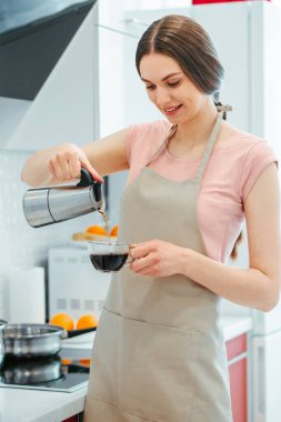 Önlüklü gülümseyen genç bayan cam bardak tutuyor ve içine kahve döküyor.