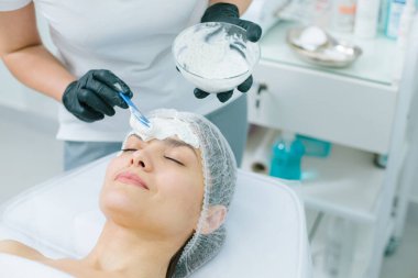 Bir kozmetikçi alnına aljinat maskesi takarken sakin bir kadın gözlerini kapatıyor.