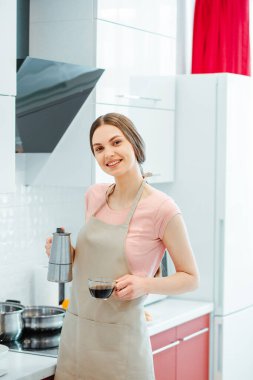Mutfakta elinde kahve demliği ve elinde bardakla bekleyen güzel, neşeli bir kadın.