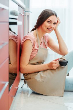 Elinde bir fincan kahveyle fırının yanında oturan önlüklü neşeli bayan.