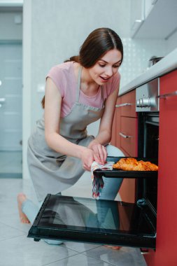 Fırını açarken gülümseyen genç bir kadın ve taze kruvasanlara bakıyor.