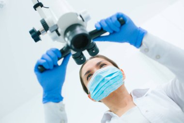 Diş mikroskobu kullanırken plastik eldiven ve tıbbi maske takan profesyonel bir kadın doktor.