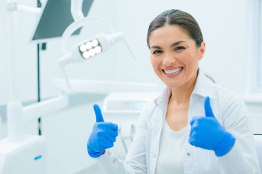 Beyaz önlüklü ve lastik eldivenli tutkulu kadın doktor klinikte iki parmağını kaldırıp gülümsüyor.