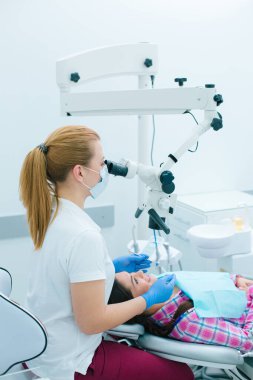 Deneyimli kadın dişçi kök kanallarını tedavi ederken hastasının dişlerine mikroskopla bakıyor.