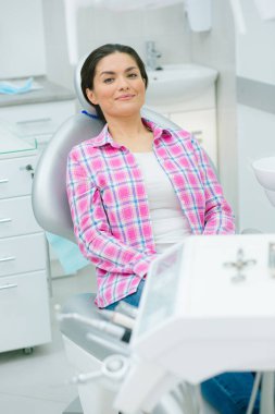 Kendine güveni tam genç bir kadın bir diş kliniğinin tıbbi koltuğunda oturuyor ve gülümsüyor.