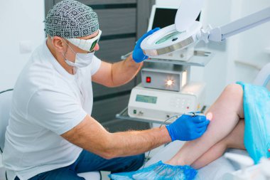 Profesyonel ekipman ve büyüteç kullanarak hastasının bacağındaki damarları tedavi eden konsantre bir dermatolog.