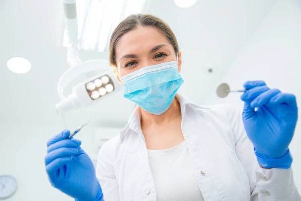 Female dentist in a medical mask and rubber gloves holding the tools while standing under the lamp