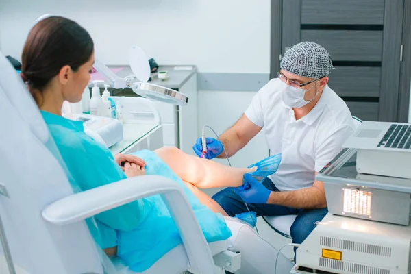 Hastasının bacağındaki damarları iyileştirirken modern ekipman kullanan tıbbi maskeli profesyonel bir doktor.