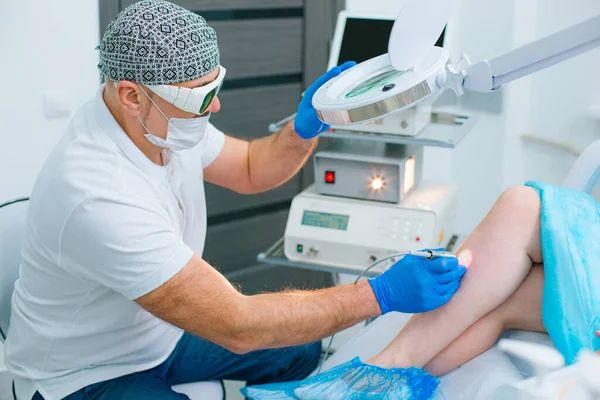 Profesyonel ekipman ve büyüteç kullanarak hastasının bacağındaki damarları tedavi eden konsantre bir dermatolog.