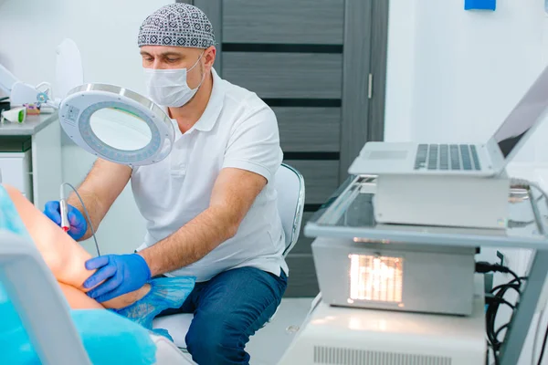 Deneyimli bir doktor, varis damarlarını lazerle tedavi ederken hastanın bacağındaki büyüteçten bakıyor.