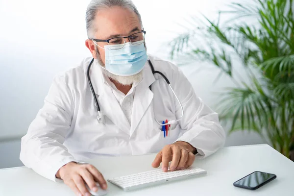 Medico in cappotto bianco con maschera viso che lavora telematicamente nel suo ufficio — Foto Stock