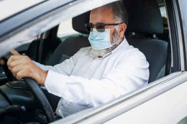 Tıbbi maske takmış araba kullanan kıdemli bir adam. Coronavirüs salgını konsepti.