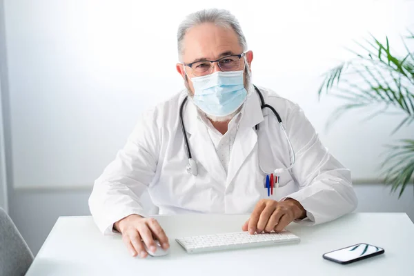 Dokter in witte jas met gezichtsmasker kijkend naar camera terwijl hij telematisch in zijn kantoor werkt — Stockfoto