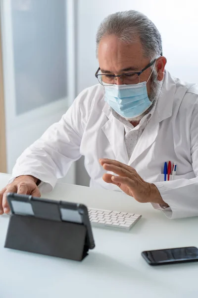 Medico in cappotto bianco con maschera facciale saluto in videoconferenza in un tablet nel suo ufficio — Foto Stock