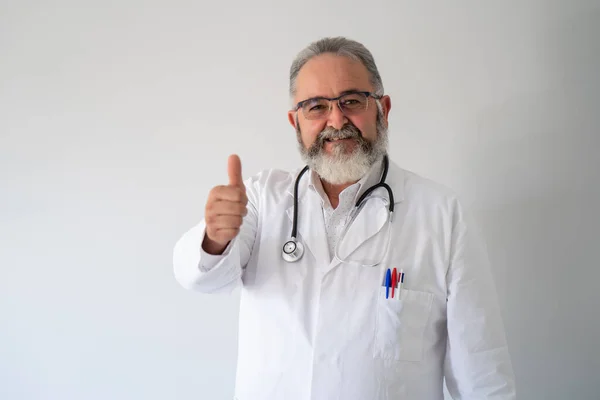 Retrato del médico barbudo sénior en bata blanca sonriendo con los pulgares hacia arriba sobre fondo blanco. — Foto de Stock