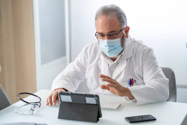 Medico in cappotto bianco con maschera facciale avendo una videoconferenza con tablet nel suo ufficio — Foto Stock