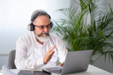 Kulaklıklı yaşlı adam video konferansında evde dizüstü bilgisayarla selamlaşıyor.