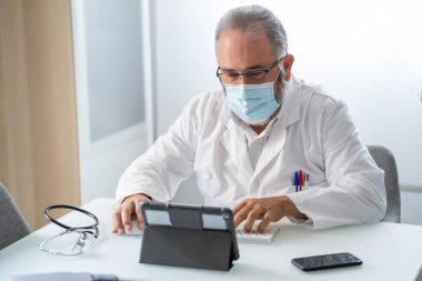 Yüz maskeli doktor ofisinde tabletle video konferansı yapıyor. 