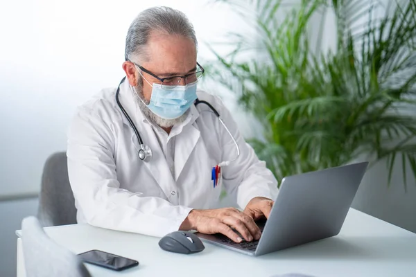 Un dottore anziano che lavora con un portatile nel suo ufficio. Concetto di lavoro telematico o online. — Foto Stock