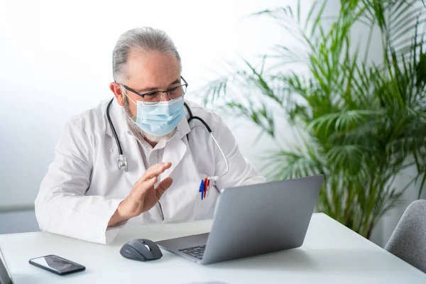 Un dottore anziano che saluta in videoconferenza un portatile con un paziente. Concetto di lavoro telematico o online. — Foto Stock