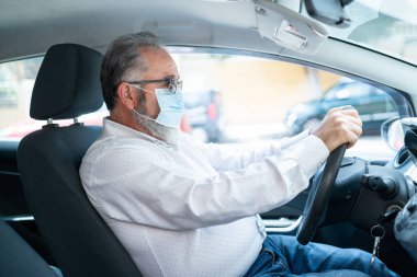 Tıbbi maske takan yaşlı bir adamın araba sürüşünün yan görüntüsü. Coronavirüs salgını konsepti.