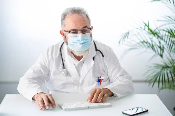 Dokter in witte jas met gezichtsmasker tijdens het werken telematisch in zijn kantoor — Stockfoto