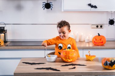 Cadılar Bayramı balkabağı tişörtü giymiş bir çocuk Cadılar Bayramı balkabağını evdeki mutfakta boşaltıyor ve dekore ediyor.. 