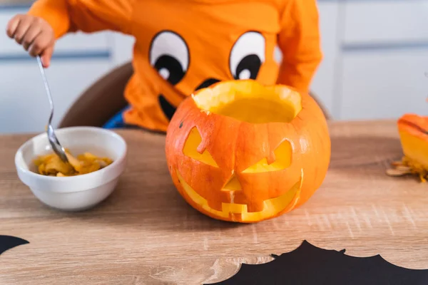 Primo piano di un bambino vestito da zucca di Halloween che svuota una zucca di Halloween nella sua cucina a casa. — Foto Stock