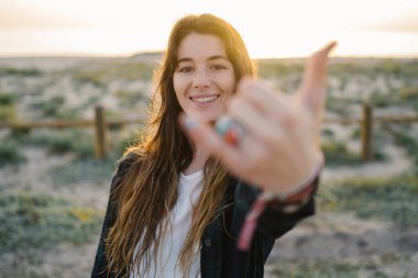 Genç bir kadın gülümsüyor ve altın saatinde kumlu bir kumsalda Shaka jesti yapıyor.